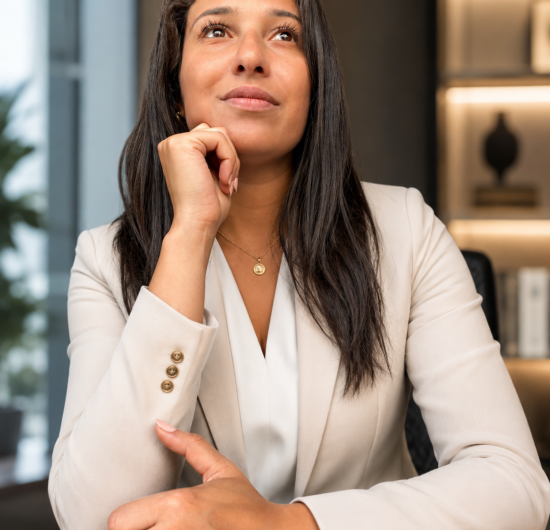 Portrait professionnel d’une femme assise à un bureau, en posture réfléchie, avec la main posée sous le menton dans un environnement corporate.